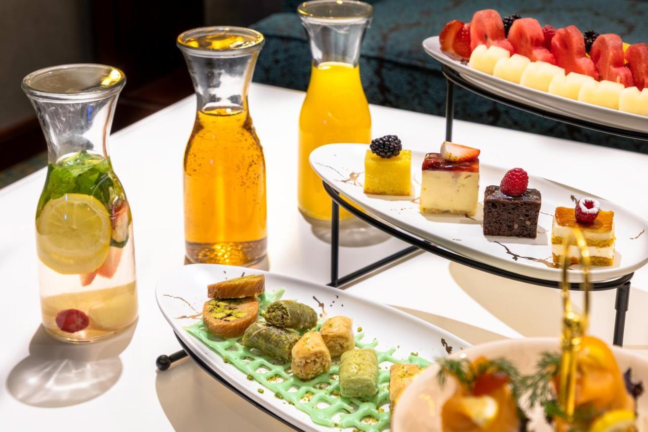 Al Aqeeq Madinah Hotel Medína Exteriér fotografie The photo showcases an elegant display of refreshments and desserts. There are several glass pitchers filled with bright, colorful beverages, likely juices or infused water, one featuring lemon and mint. A two-tier serving stand holds an assortment o