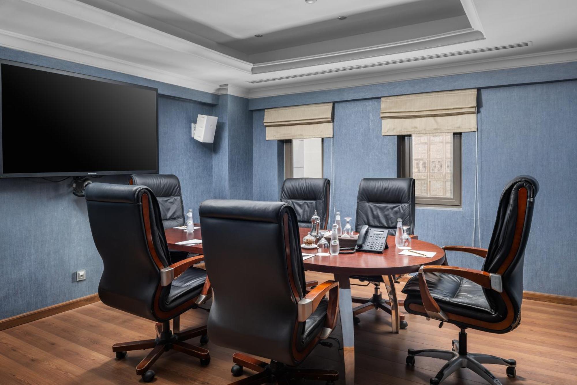 Al Aqeeq Madinah Hotel Medína Exteriér fotografie The photo shows a modern conference room. It features a round wooden table surrounded by several black leather swivel chairs. The walls have a blue color scheme, and there is a large flat-screen TV mounted on one wall. The room has two windows with b