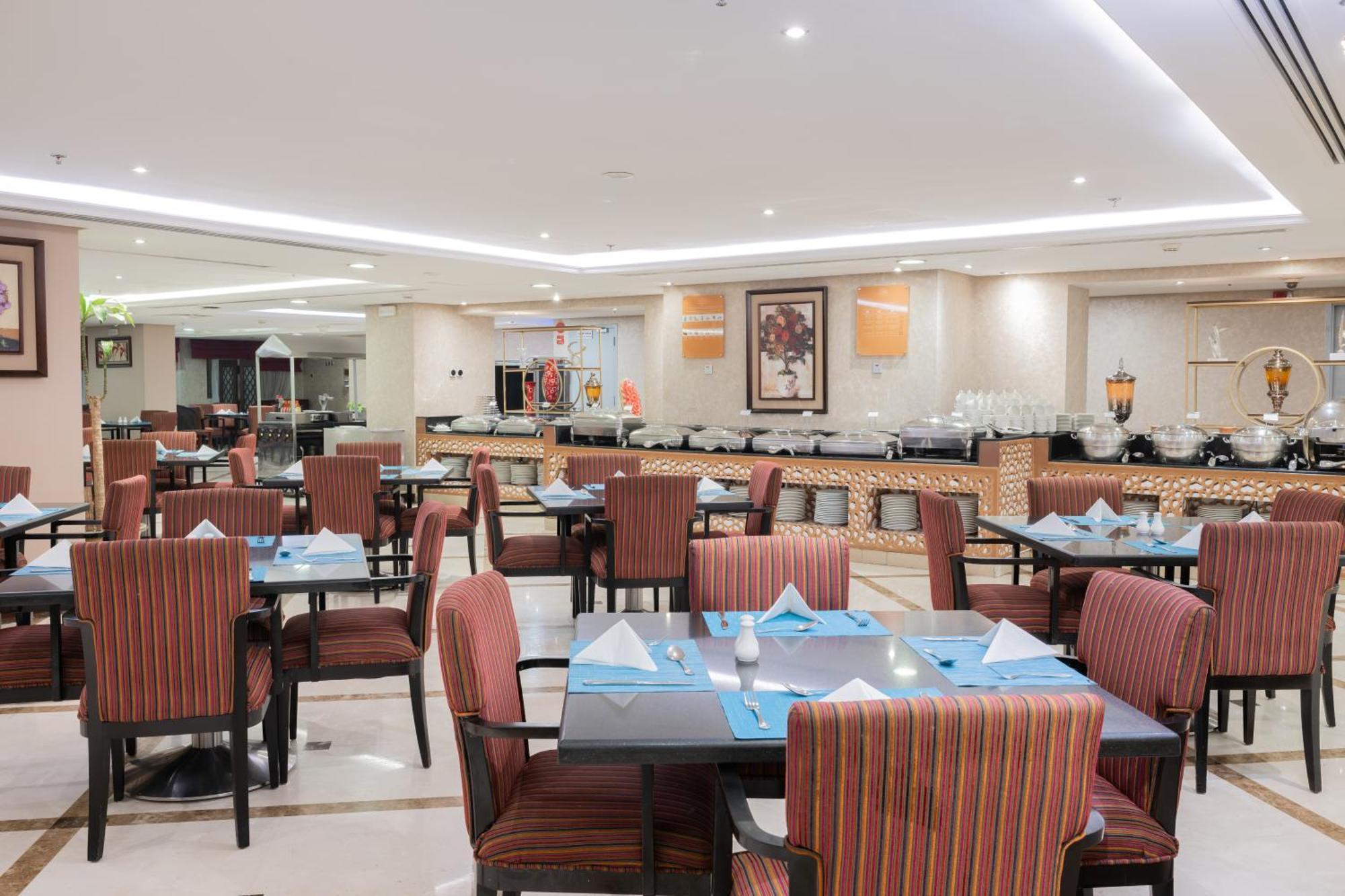 Al Aqeeq Madinah Hotel Medína Exteriér fotografie The photo shows the interior of a restaurant. It features multiple tables arranged neatly, each with patterned chairs and set with blue and white table settings, including folded napkins. The lighting is bright and modern, creating a welcoming atmosp