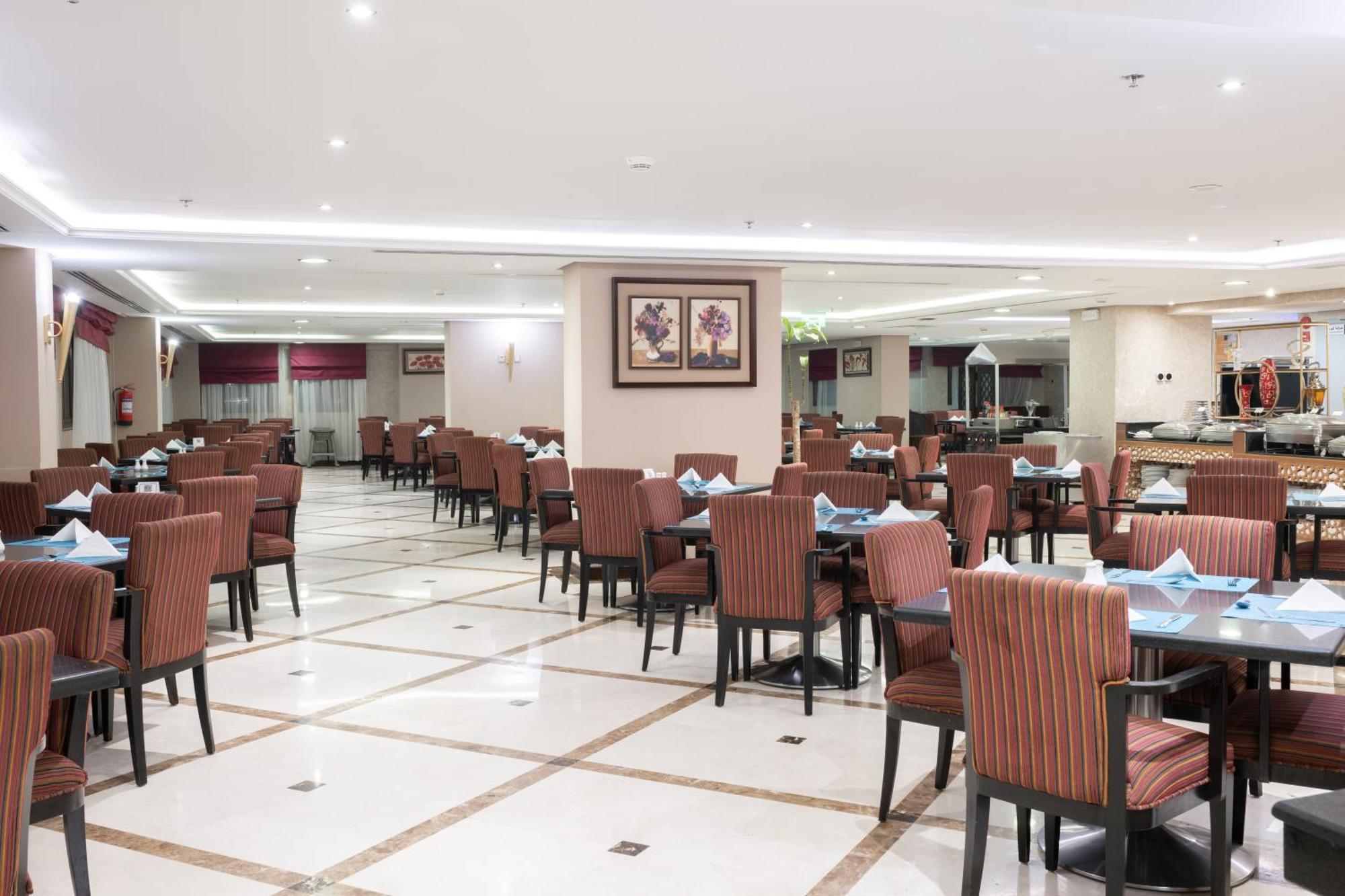 Al Aqeeq Madinah Hotel Medína Exteriér fotografie The photo shows the interior of a spacious restaurant. It features a well-organized dining area with neatly arranged tables and chairs. The tables are dressed with blue napkins, and there is a light-colored flooring with a grid pattern. The walls are