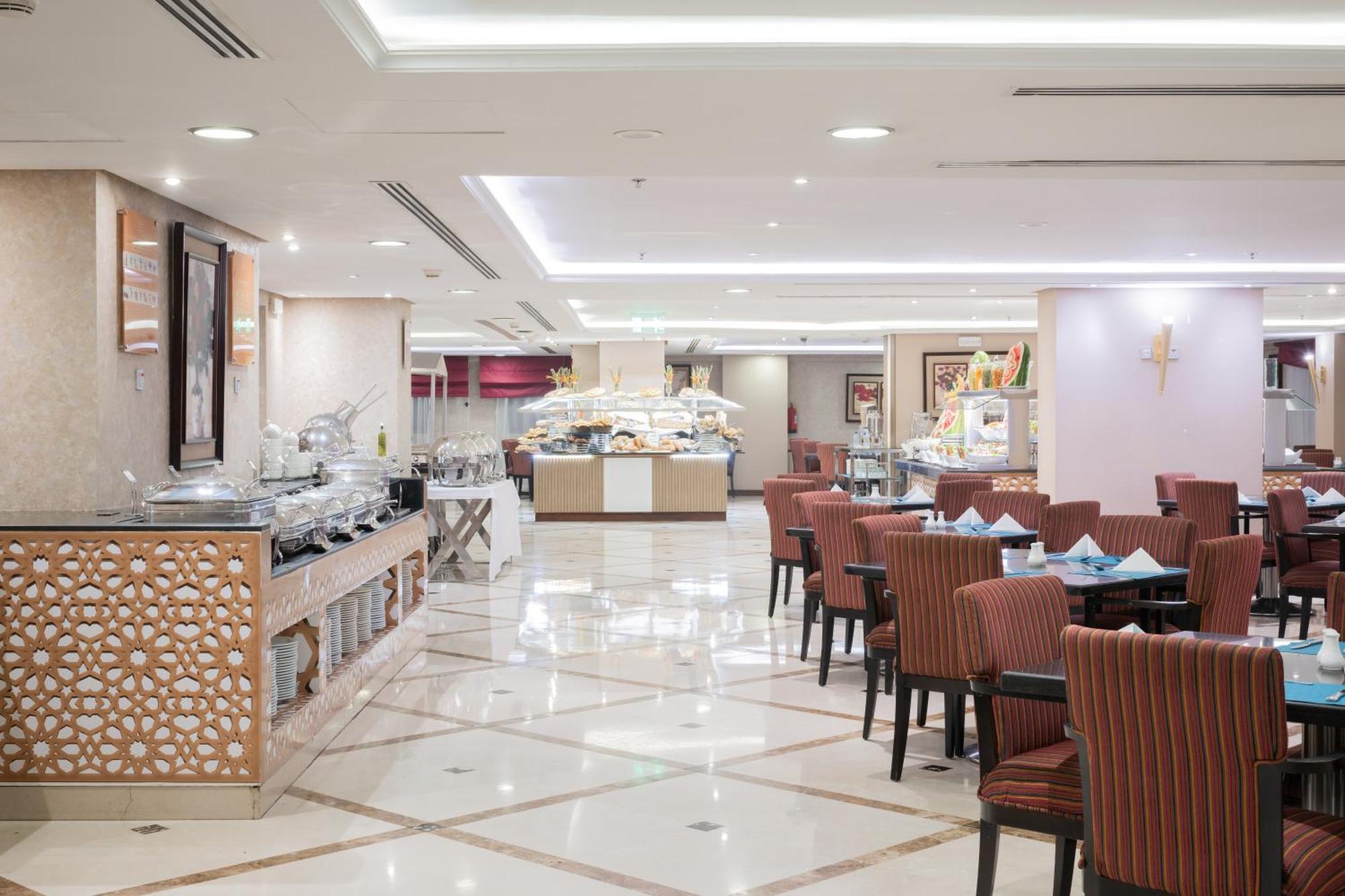 Al Aqeeq Madinah Hotel Medína Exteriér fotografie The photo shows an elegantly designed restaurant or dining area. It features a spacious layout with a glossy tiled floor and bright, soft lighting. There are several dining tables with striped chairs arranged neatly. Along one side, there's a buffet 