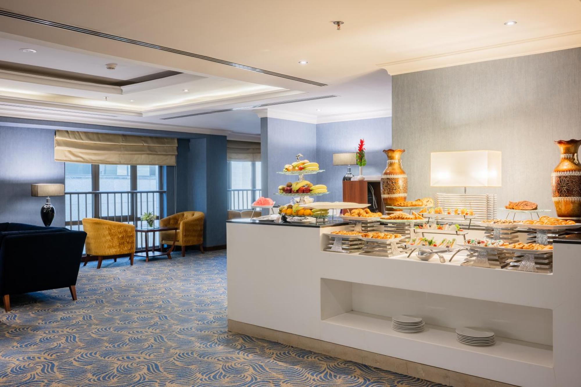 Al Aqeeq Madinah Hotel Medína Exteriér fotografie The photo shows a modern dining area, likely in a hotel or restaurant. On the right side, there is a buffet table with various food items displayed, including pastries and desserts. Behind the table, a decorative fountain is visible. The room feature