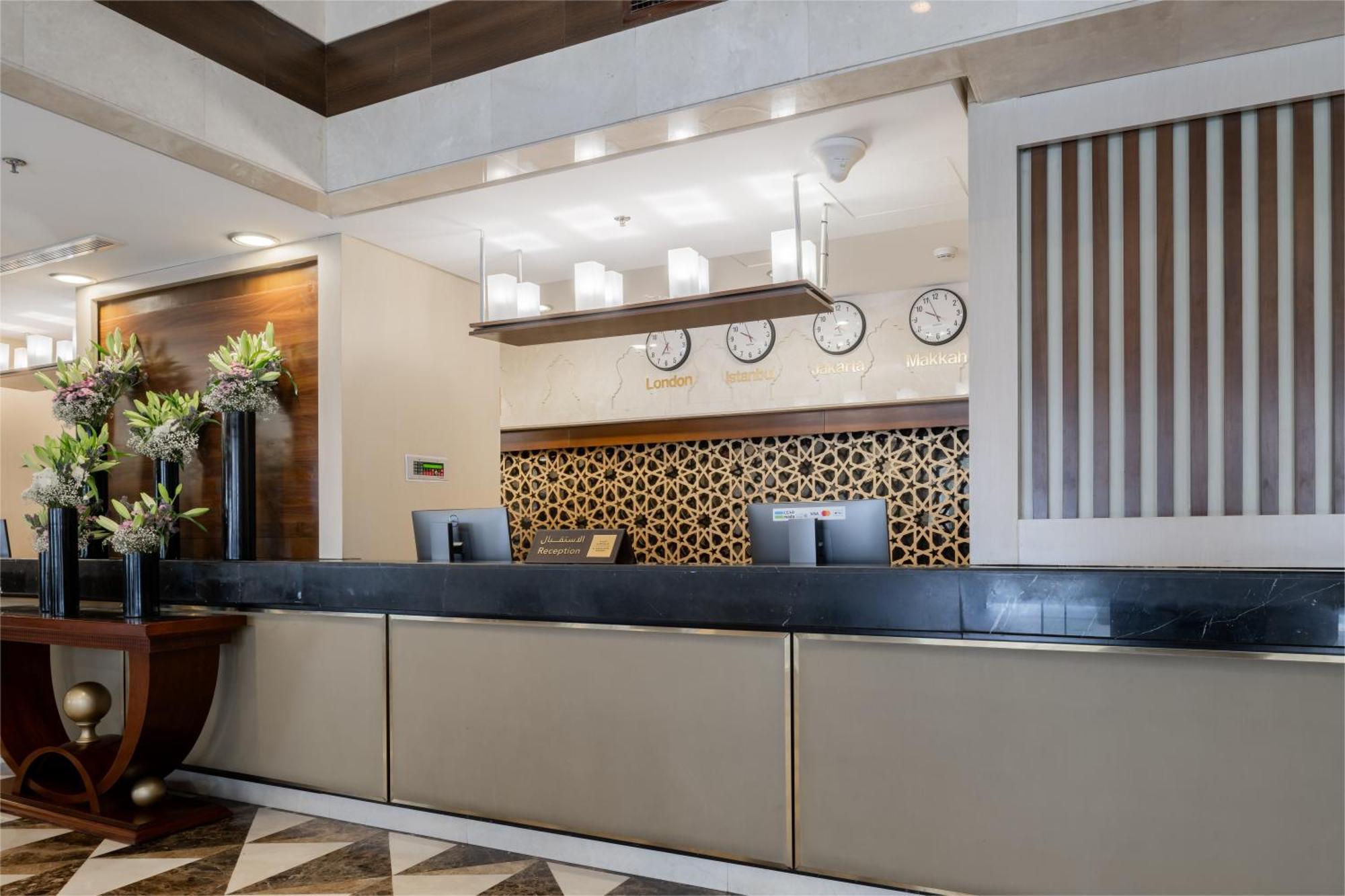 Al Aqeeq Madinah Hotel Medína Exteriér fotografie The photo shows the interior of a hotel reception area. The reception desk is designed with a sleek, modern look featuring a dark countertop and a light-colored front. Above the desk, there are several clocks displaying different times, suggesting an