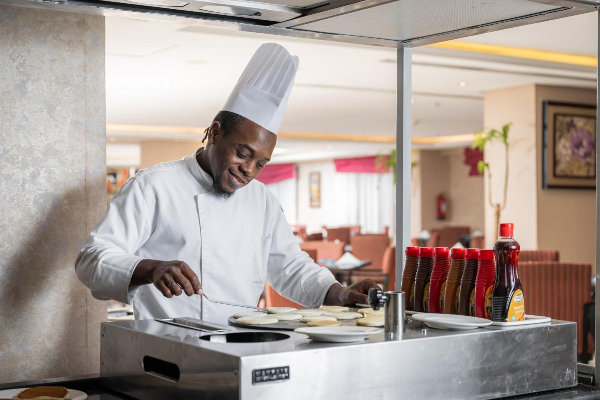 Al Aqeeq Madinah Hotel Medína Exteriér fotografie The photo shows a chef in a professional kitchen setting. He is wearing a traditional white chef's uniform and a tall white chef's hat, and he appears to be smiling while cooking. The chef is standing at a stainless steel cooking station, where he is