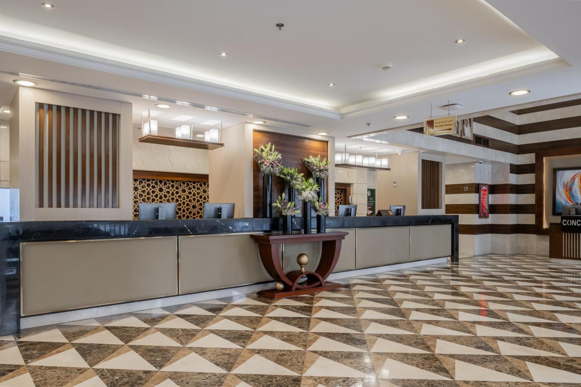 Al Aqeeq Madinah Hotel Medína Exteriér fotografie The photo depicts a hotel reception area. The check-in counter is long and sleek, with a smooth surface and decorative elements. Behind the counter, there are several computer monitors. The walls feature stylish designs, including wooden panels and g