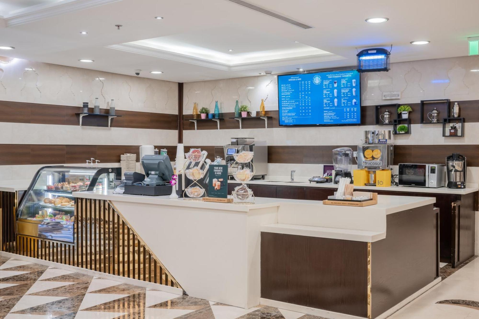 Al Aqeeq Madinah Hotel Medína Exteriér fotografie The photo shows a modern café or coffee shop interior. There's a counter area made of smooth white and dark brown materials, featuring a cash register and display items. Behind the counter, shelves are stocked with various beverage items and equipmen