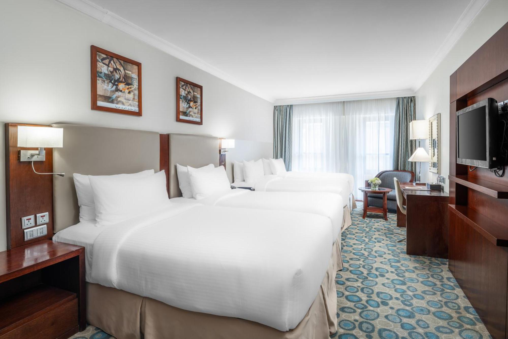 Al Aqeeq Madinah Hotel Medína Exteriér fotografie The photo shows a hotel room featuring a modern, clean design. There are two large beds with white linens positioned side by side. Above the beds are framed artworks, and soft lighting is provided by wall sconces. To the right, there is a wooden ente