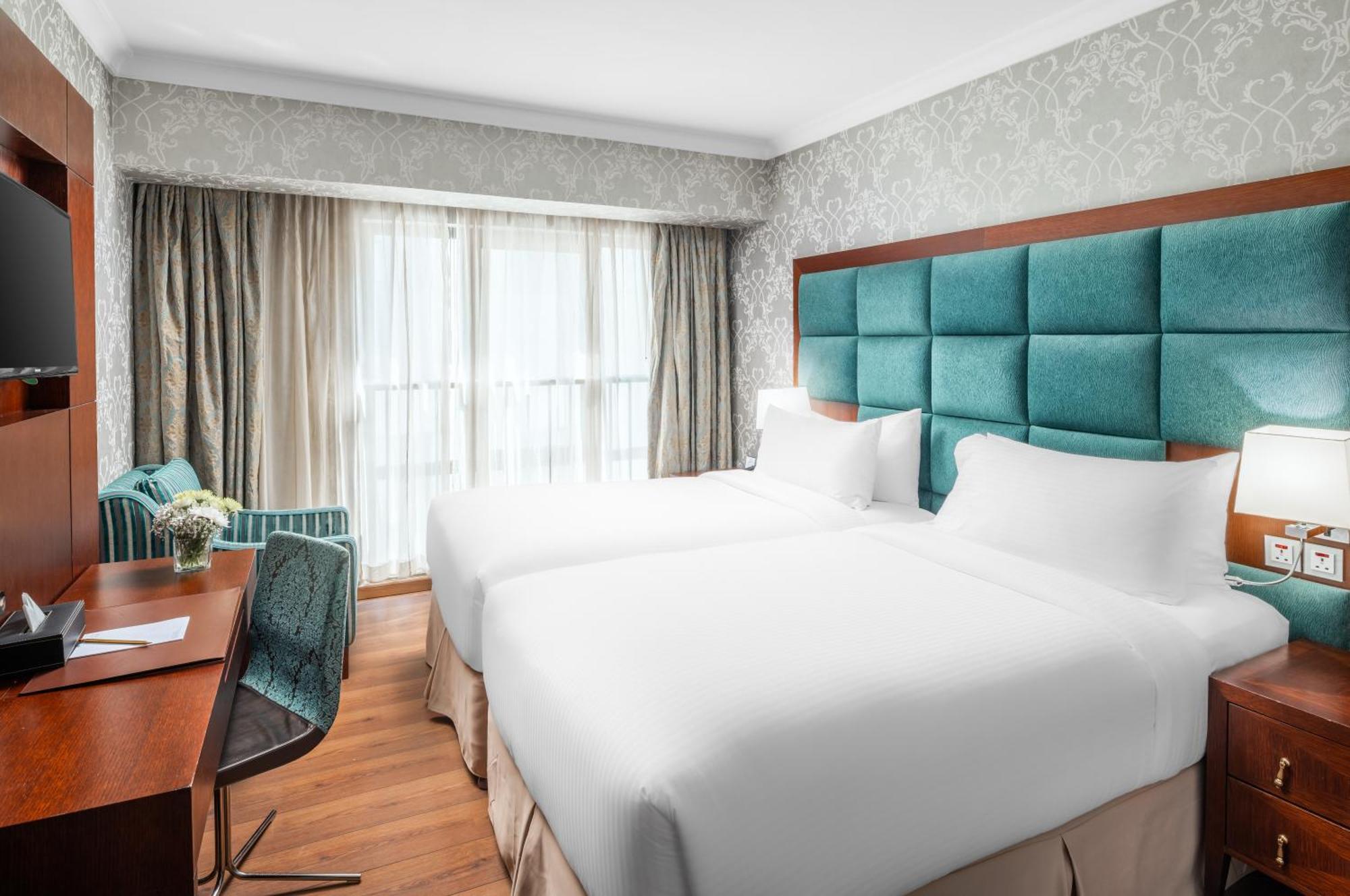 Al Aqeeq Madinah Hotel Medína Exteriér fotografie The photo shows a modern hotel room featuring two single beds with white bedding. The headboard is upholstered in a teal fabric. There is a wooden desk and a chair near a window that is covered with sheer curtains and drapes. A cozy armchair in a mat