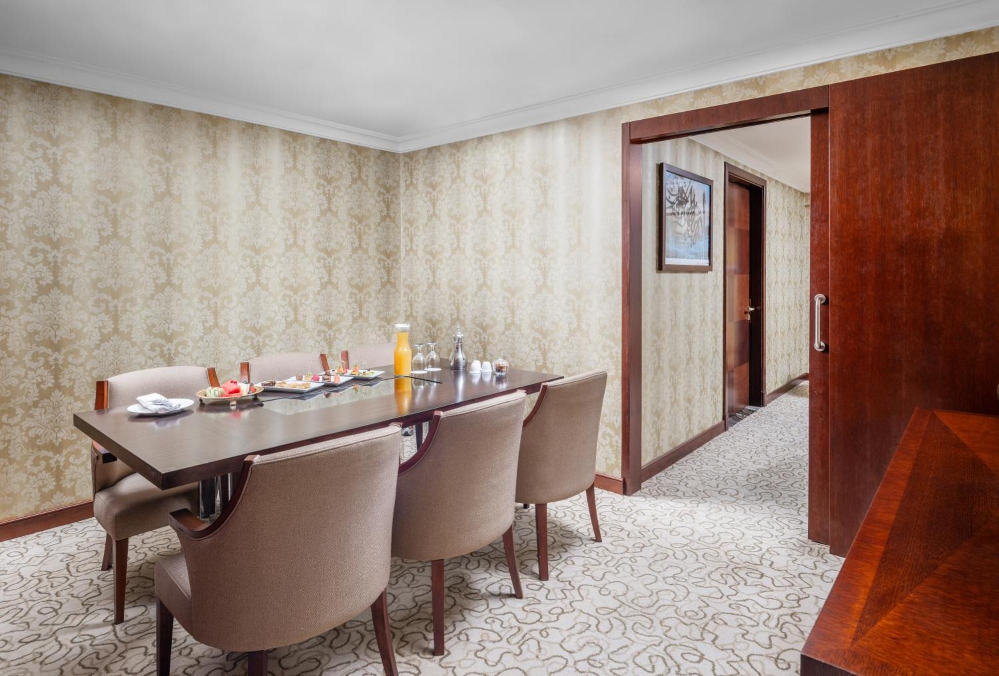 Al Aqeeq Madinah Hotel Medína Exteriér fotografie The photo shows a dining room with a stylish and elegant design. There is a large wooden dining table surrounded by plush, beige chairs. On the table, there are various items including a bottle of orange juice, glasses, and other dining accessories. 