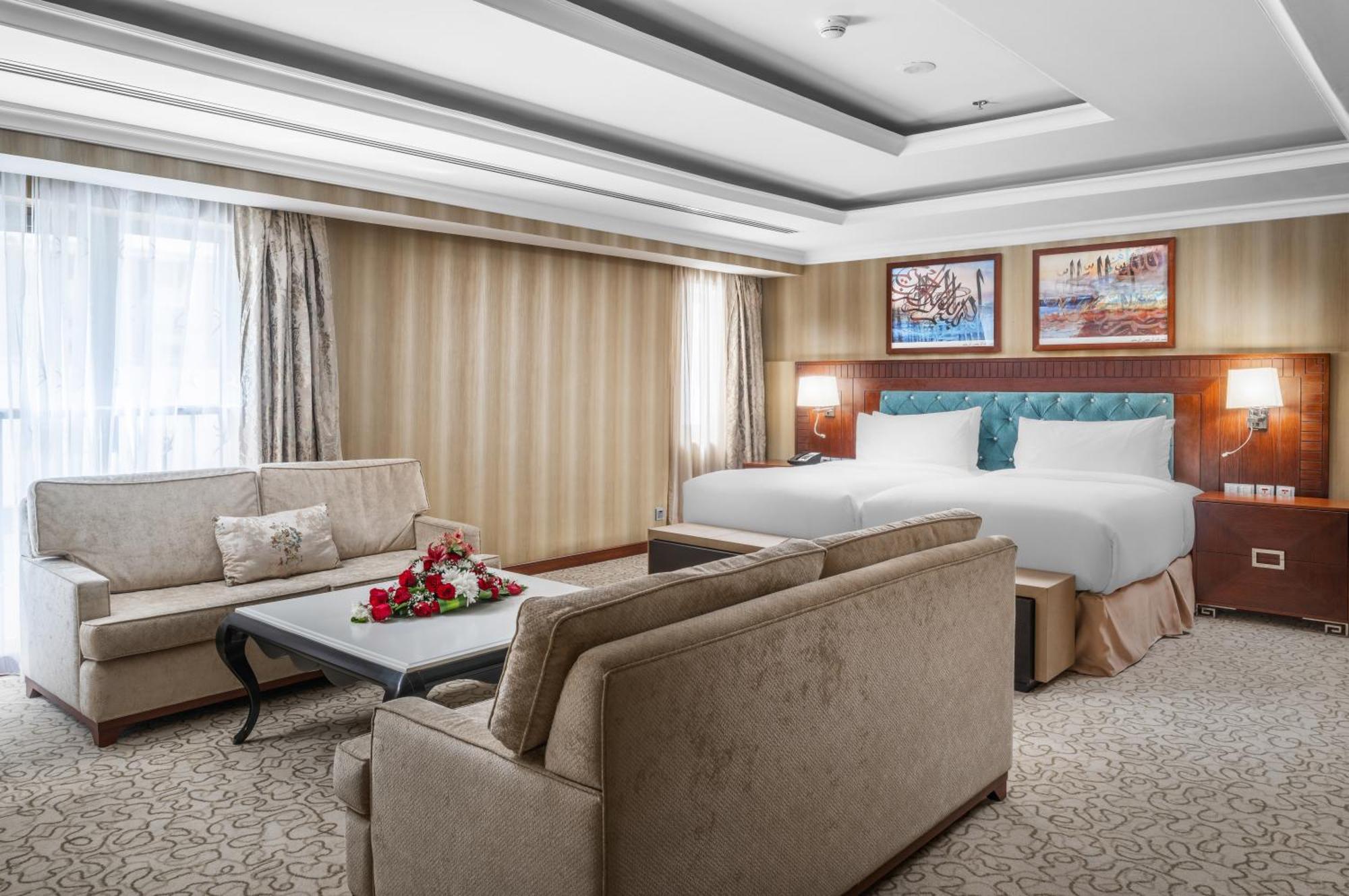 Al Aqeeq Madinah Hotel Medína Exteriér fotografie The photo shows a well-decorated hotel room. It features a large bed with two pillows and a light-colored duvet. There are two comfortable couches arranged around a coffee table with a floral centerpiece. The walls have a subtle striped design, and l