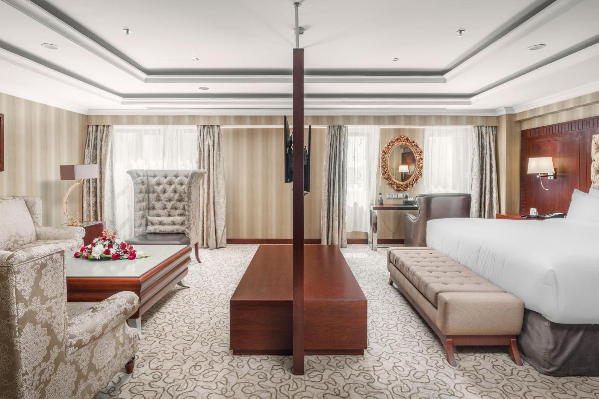 Al Aqeeq Madinah Hotel Medína Exteriér fotografie The photo showcases a spacious hotel suite with an elegant and modern design. The room features a large bed on one side, complemented by a plush bench at the foot. Opposite the bed, there is a stylish seating area that includes a decorative armchair 