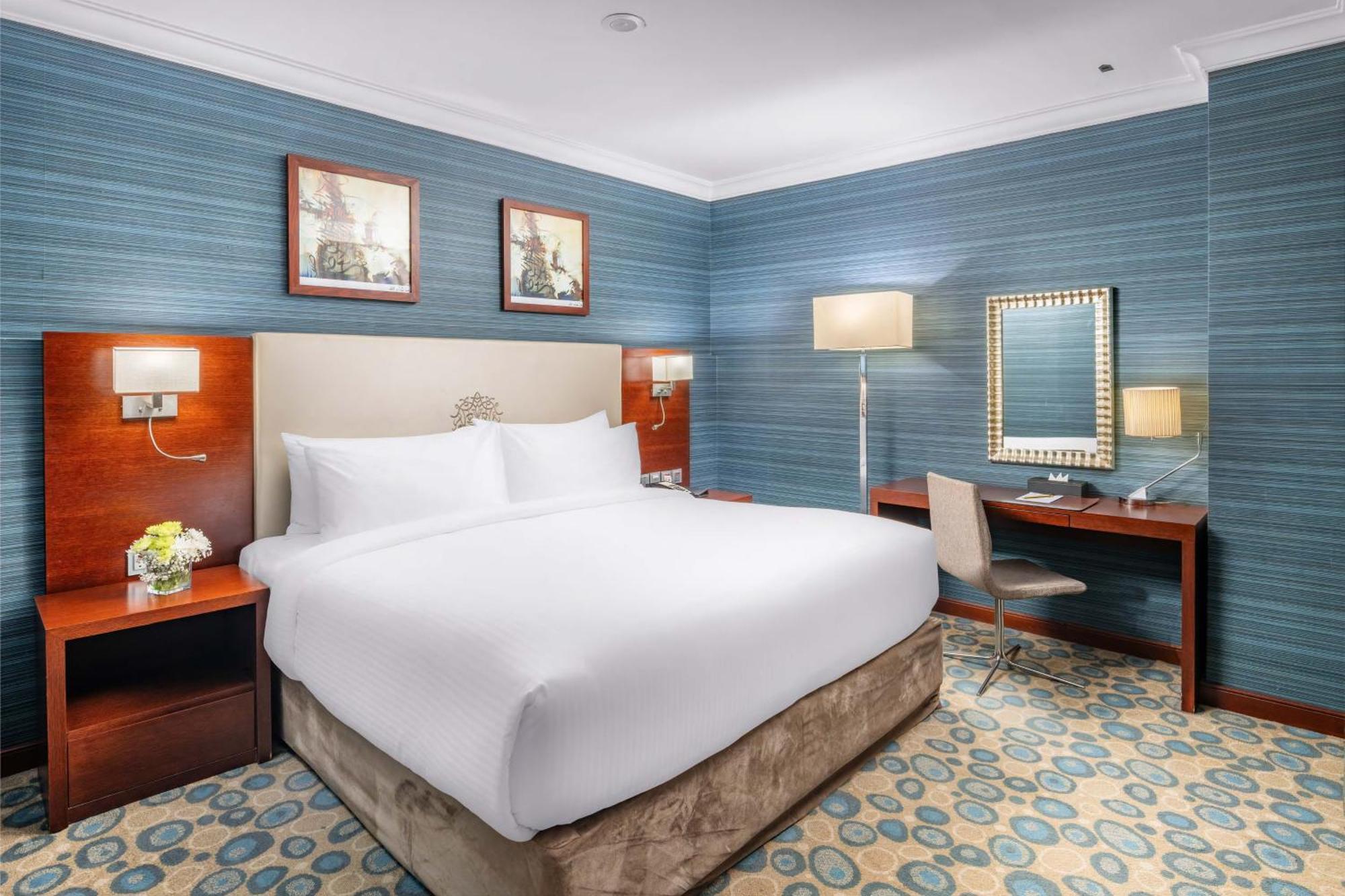 Al Aqeeq Madinah Hotel Medína Exteriér fotografie The photo shows a well-furnished hotel room. It features a large bed with white linens, flanked by wooden nightstands. The walls are painted in a light blue color with a textured pattern. Above the bed, there are two framed pictures. On one side of t