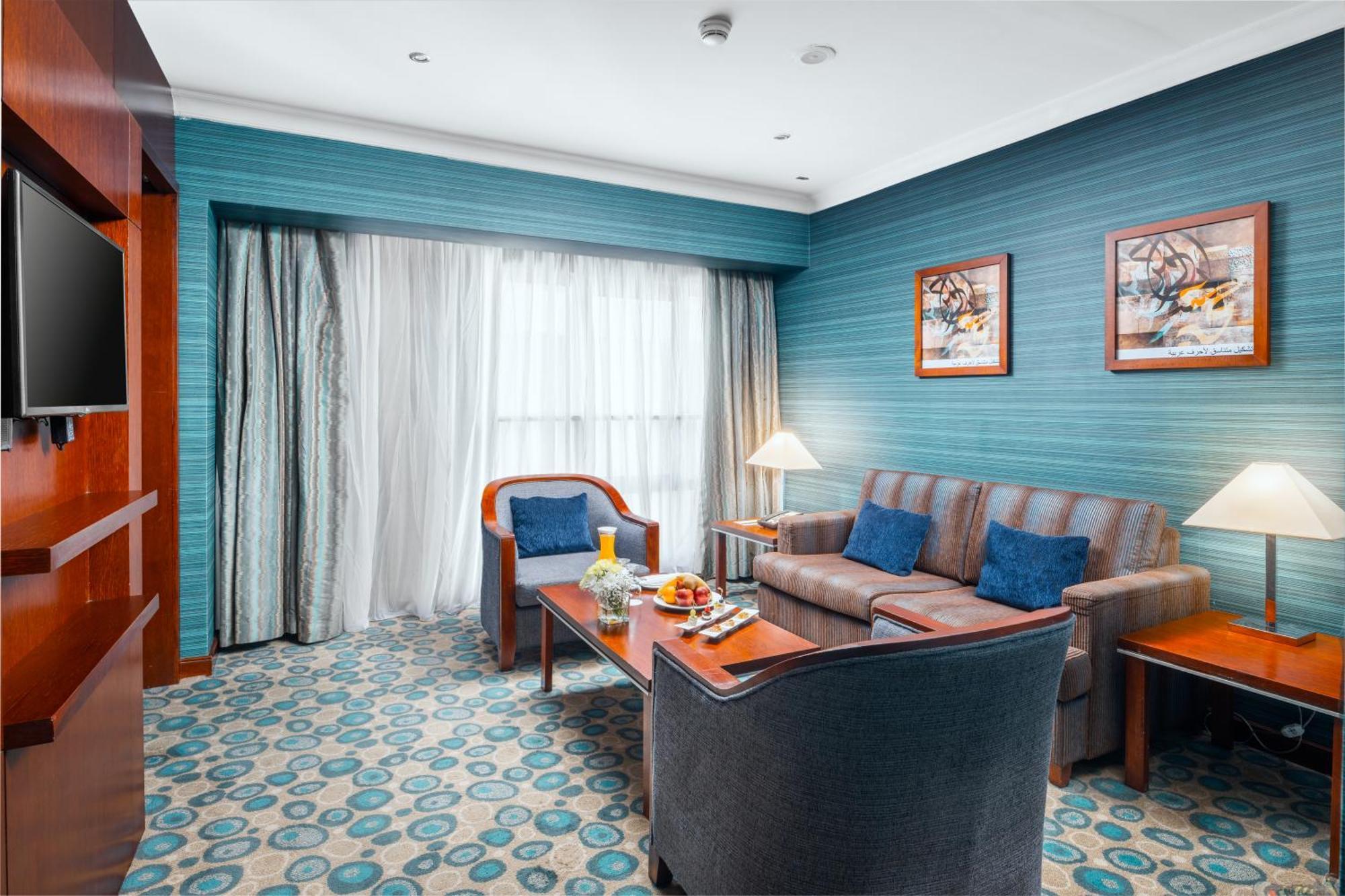 Al Aqeeq Madinah Hotel Medína Exteriér fotografie The photo shows a cozy and modern hotel room featuring a sitting area. The walls are painted a calming teal color, complemented by a patterned carpet in shades of blue and beige. There is a comfortable sofa with cushions, two armchairs, and a wooden 