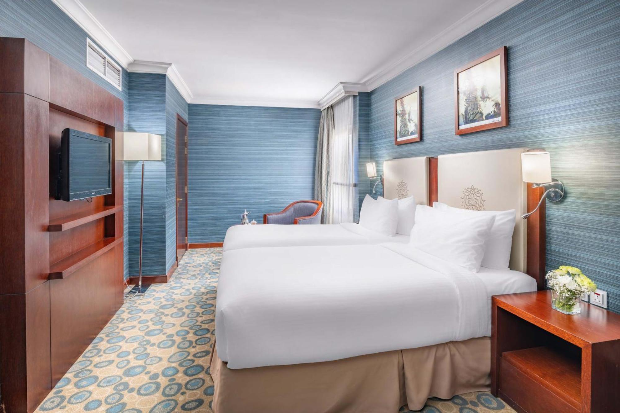 Al Aqeeq Madinah Hotel Medína Exteriér fotografie The photo shows a hotel room featuring two twin beds dressed in white linens. The walls are painted in a soft blue color, and there are framed artworks hung above the beds. A flat-screen TV is mounted on a wooden unit against one wall. To the side, t