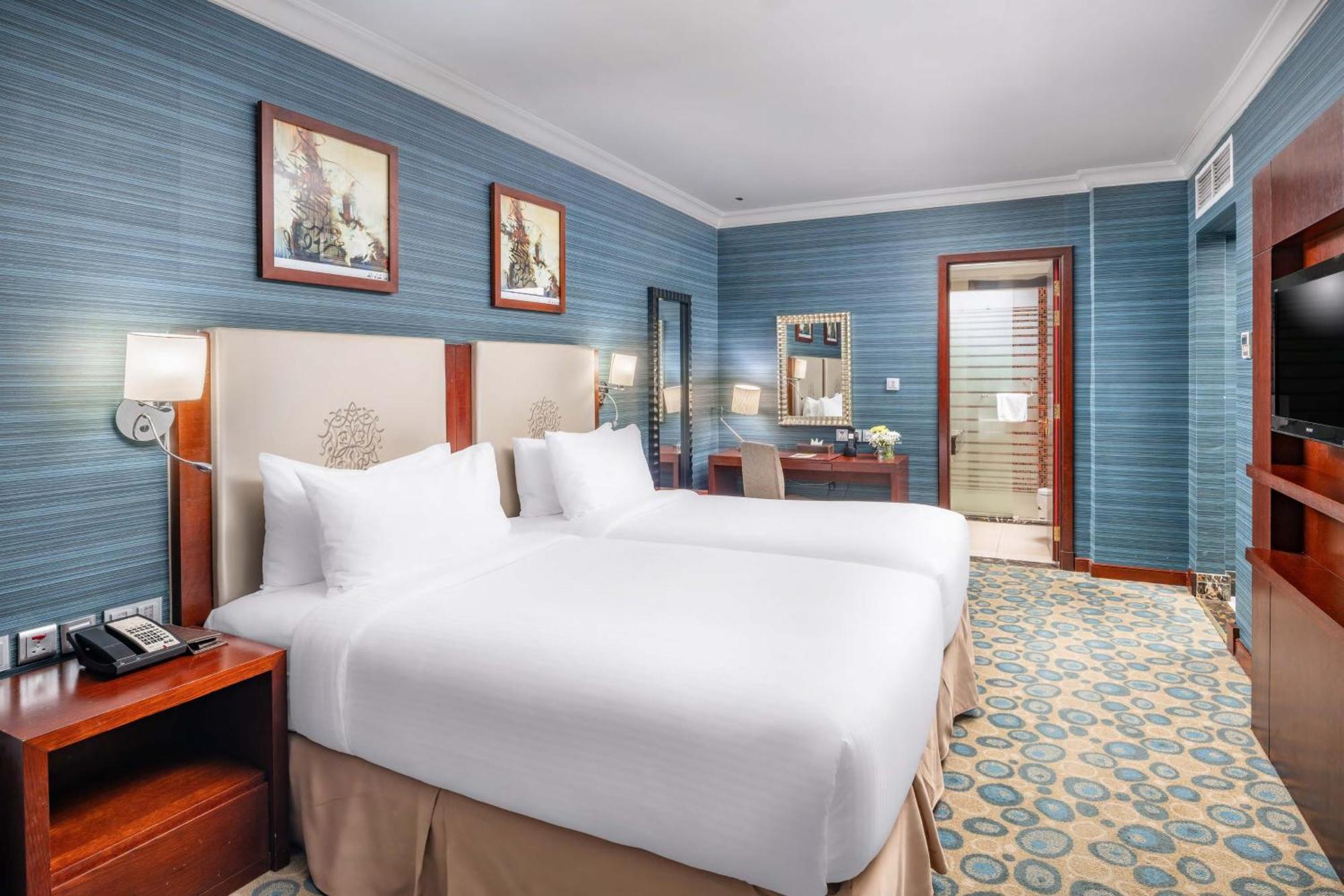 Al Aqeeq Madinah Hotel Medína Exteriér fotografie The photo shows a hotel room featuring two twin beds with white bedding, positioned against a light-colored headboard. The walls are painted in a soft blue shade, adorned with framed artwork. There is a wooden desk with a chair, and a mirror mounted 