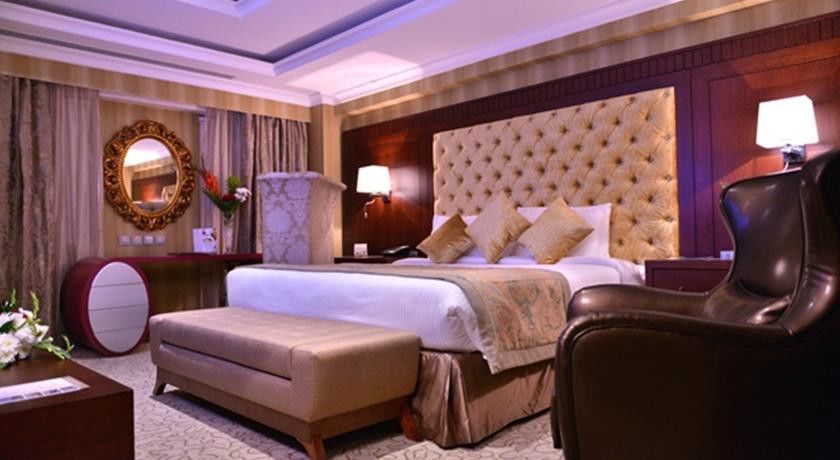Al Aqeeq Madinah Hotel Medína Exteriér fotografie The photo shows a luxurious hotel room. It features a large, upholstered headboard with a tufted design, paired with plush bedding and decorative cushions. There's a stylish bench at the foot of the bed and a comfortable armchair to the side. The roo