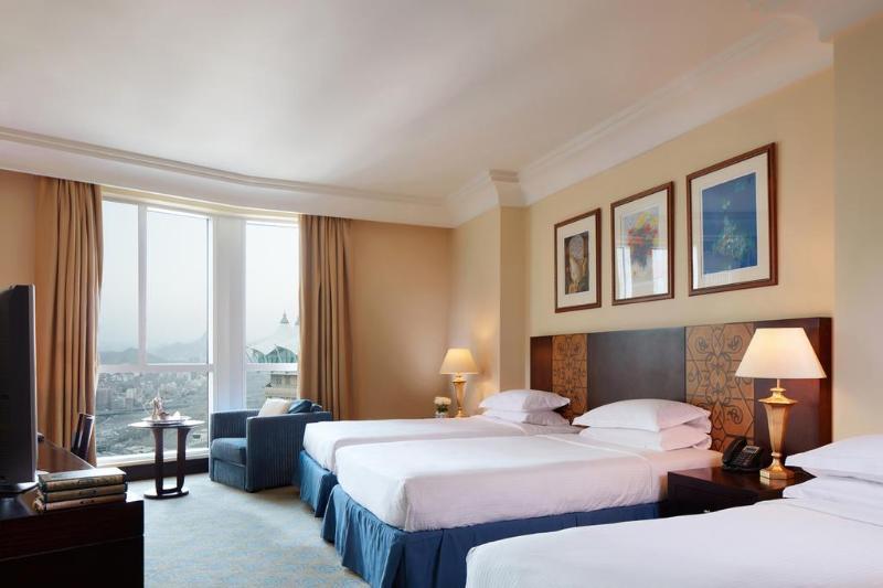 Al Aqeeq Madinah Hotel Medína Exteriér fotografie The photo shows a hotel room featuring three beds arranged neatly. The room has a warm, inviting ambiance with beige walls and a soft carpet. Large windows allow natural light to fill the space, offering a view outside. There are curtains in a light 