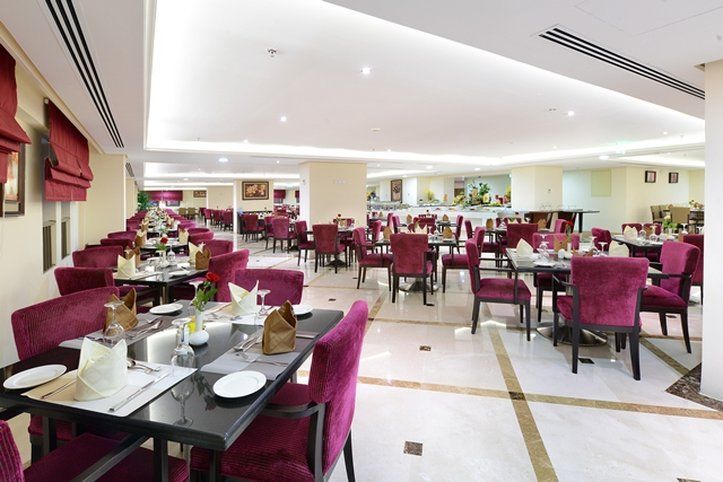 Al Aqeeq Madinah Hotel Medína Exteriér fotografie The photo shows an elegant restaurant interior with a spacious layout. The dining area features multiple tables, each set with white tablecloths and dinnerware. The chairs are upholstered in a rich purple fabric, adding a touch of luxury to the atmos
