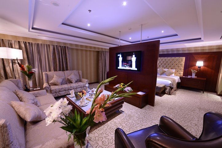 Al Aqeeq Madinah Hotel Medína Exteriér fotografie The photo shows a luxurious hotel room suite. There is a comfortable seating area with stylish sofas and a coffee table. A decorative arrangement of flowers is on the table, adding a touch of elegance. A large flat-screen television is mounted on a p