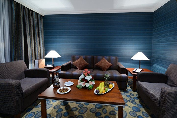 Al Aqeeq Madinah Hotel Medína Exteriér fotografie The photo shows a cozy living area featuring a dark blue wall with a textured design. In the center, there is a wooden coffee table surrounded by two comfortable armchairs and a sofa with decorative cushions. On the table, there are plates with snack