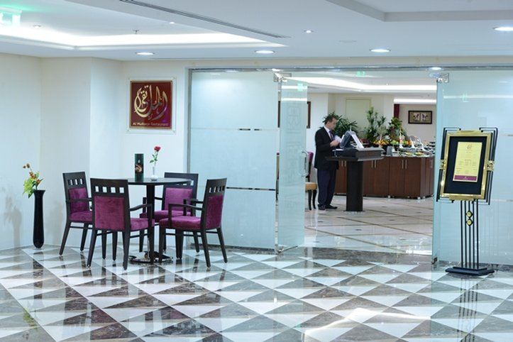 Al Aqeeq Madinah Hotel Medína Exteriér fotografie The photo shows a modern interior space, likely a lobby or reception area of a hotel or restaurant. It features a checkered floor with a combination of dark and light tiles. On the left, there is a small table set for two, adorned with a single rose 