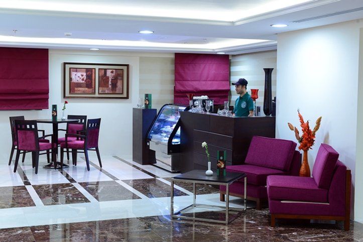 Al Aqeeq Madinah Hotel Medína Exteriér fotografie The photo shows a modern interior space, likely a café or small restaurant. It features a seating area with a few tables and chairs, all in a dark wood finish with purple upholstery. There is a counter area with a staff member behind it, possibly ser