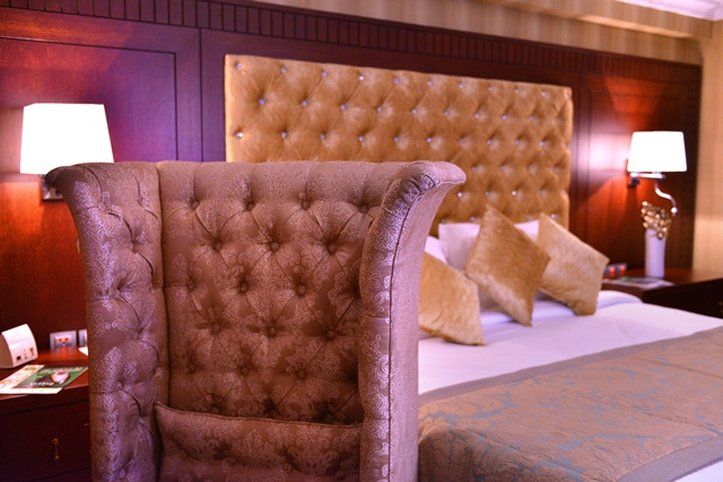 Al Aqeeq Madinah Hotel Medína Exteriér fotografie The image shows a luxurious bedroom featuring a large upholstered headboard with a tufted design in a soft, beige color. The bed is elegantly dressed with decorative pillows and a textured bedspread. In the background, there are wooden paneling and s